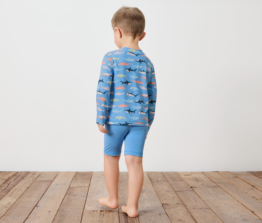 Ein Kind steht mit dem Rücken zur Kamera, es trägt ein blaues langärmliges Oberteil mit Hai-Muster und blaue Shorts.