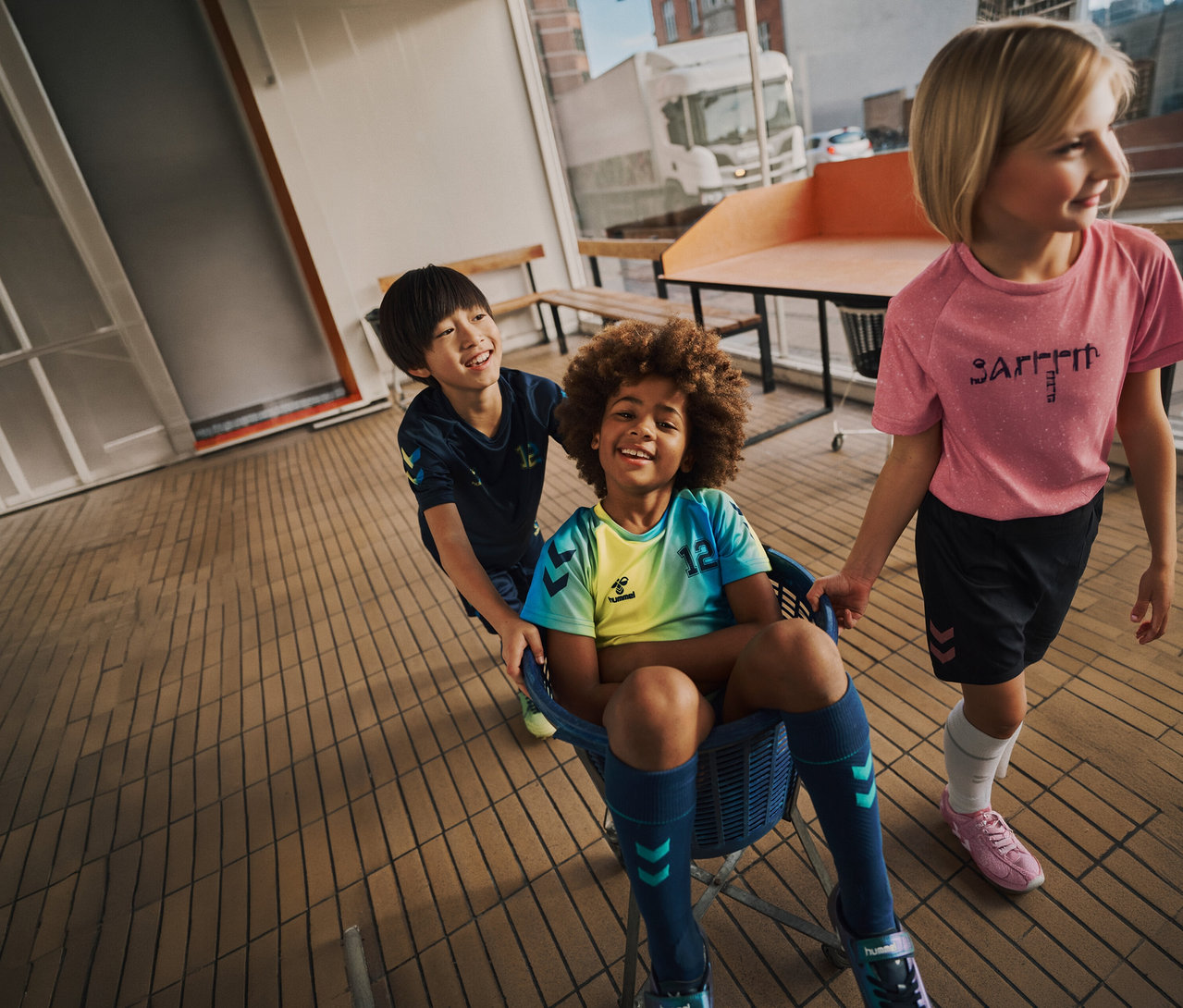 Drei Kinder, eines im Korb, tragen hummel® Kinder-Trikot-Sets HMLSHIMMER in Limette und anderen Farben.