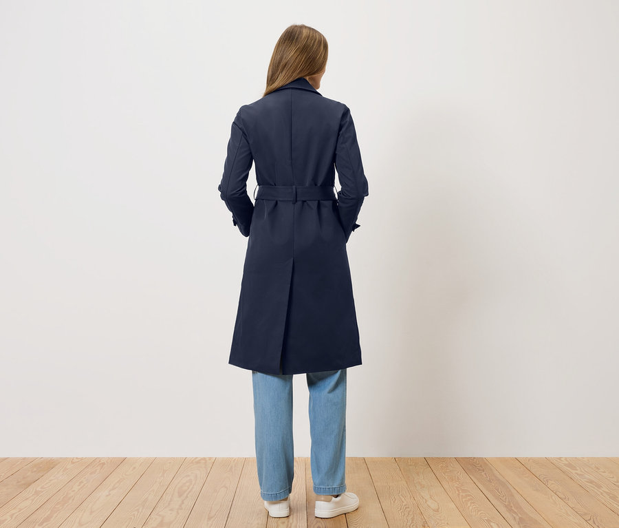 Eine Frau steht mit dem Rücken zur Kamera und trägt einen dunkelblauen Trenchcoat, Jeans und weiße Schuhe.