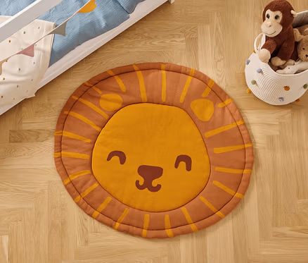 Auf einem Holzboden liegt ein runder Teppich mit einem Löwengesicht, neben einem Spielzeugkorb und einem Kinderbett.