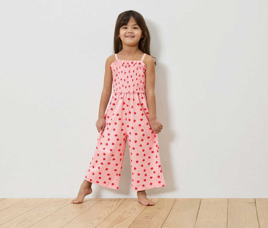Ein junges Mädchen steht barfuß vor einer weißen Wand und trägt einen rosa Overall mit Erdbeermuster.
