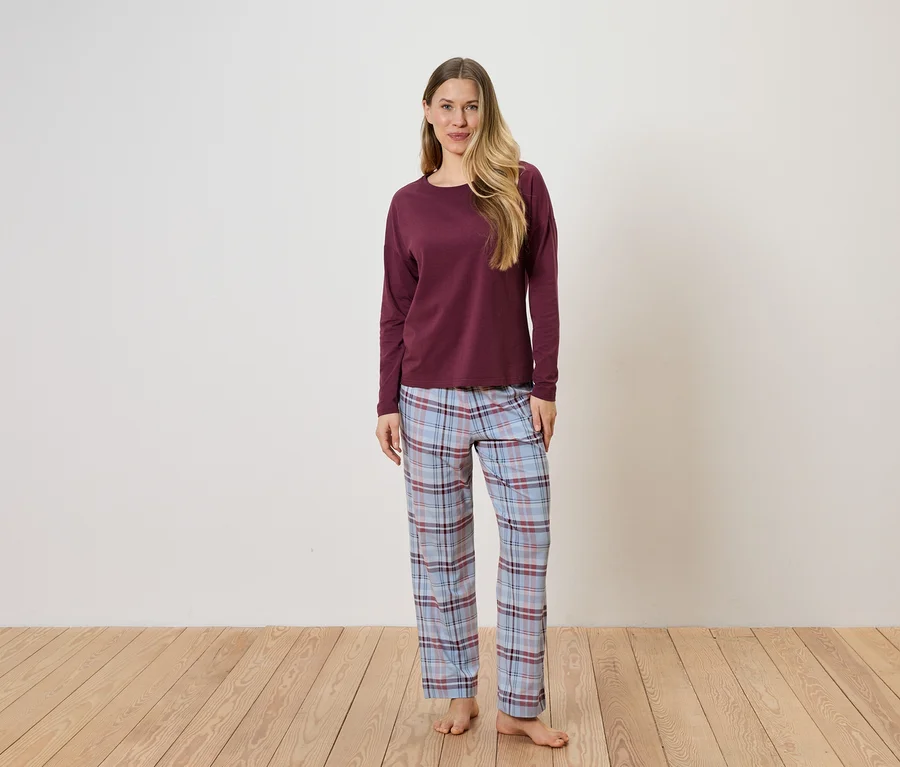 Eine Frau steht barfuß vor einer weißen Wand und trägt einen bordeauxroten Pullover und eine blau-karierte Pyjamahose.