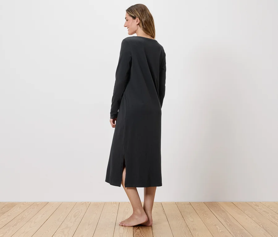 Eine Frau steht barfuß in einem langen, dunklen Kleid mit Seitenschlitz vor einer weißen Wand.