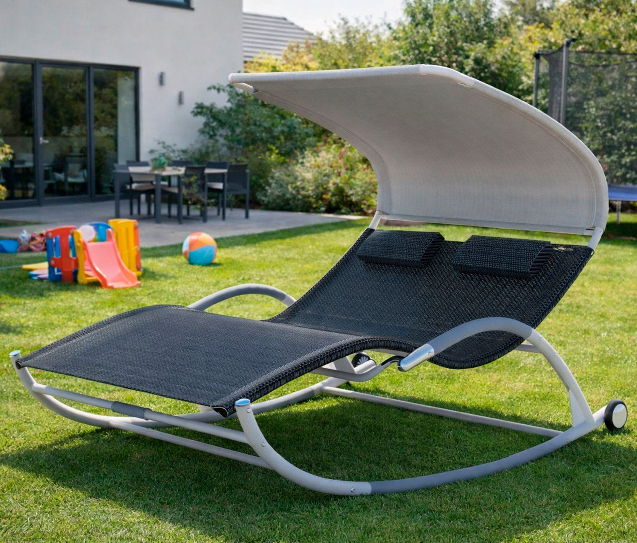 Leco Doppelschaukelliege mit Rädern auf Rasen vor Haus mit Spielplatz.
