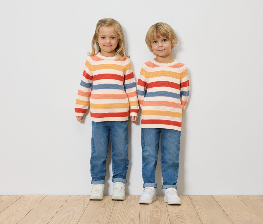 Zwei Kinder stehen nebeneinander vor einer weißen Wand. Sie tragen gestreifte Pullover, Jeans und weiße Turnschuhe.