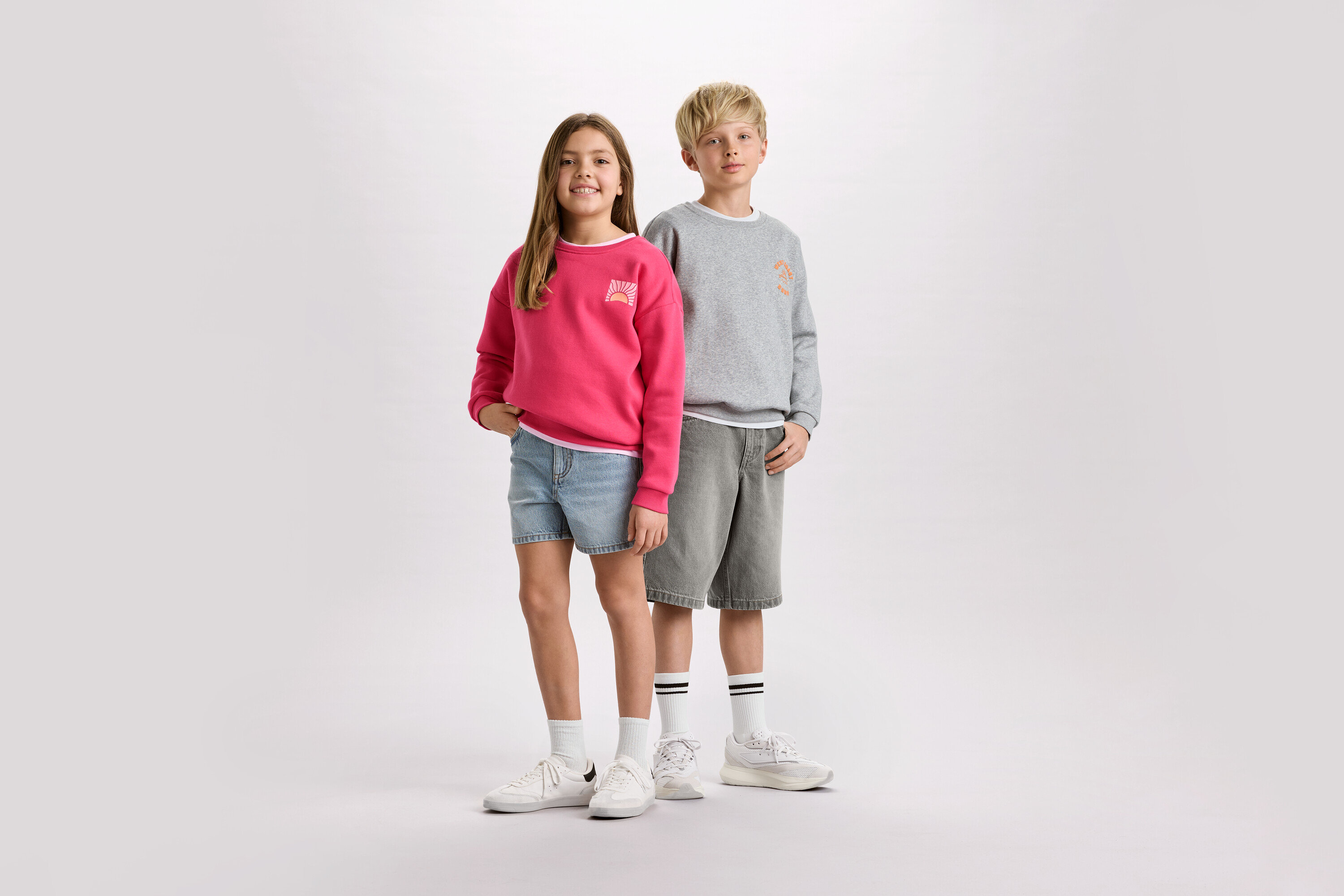 Ein Mädchen mit einem pinken Pullover und Jeansshorts steht neben einem Jungen mit einem grauen Pullover und Shorts.