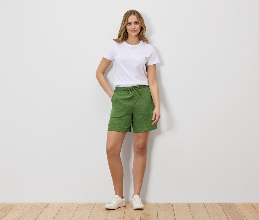 Frau in weißem T-Shirt und grünen Shorts mit Kordelzug, stehend vor einer weißen Wand.