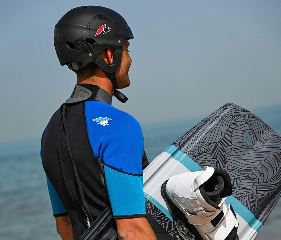 Ein Mann in einem blauen und schwarzen Neoprenanzug und einem schwarzen Helm hält ein Kiteboard vor einem Gewässer.