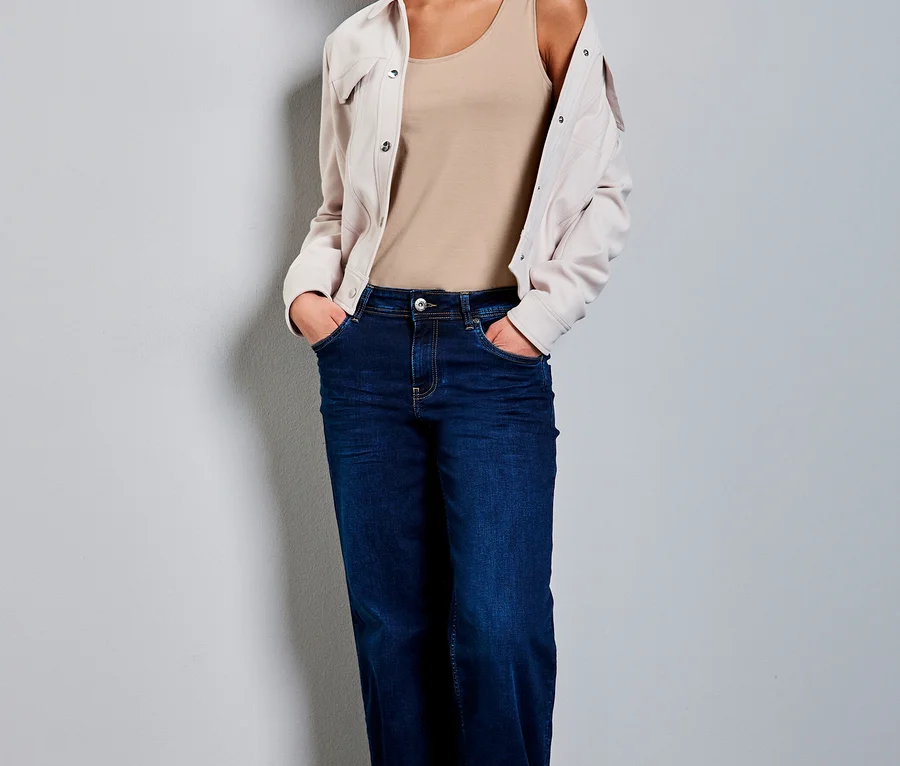 Frau trägt ein beiges Street One Basic-Top, dunkelblaue weite Jeans und eine helle Jacke über den Schultern, Hände in den Taschen.