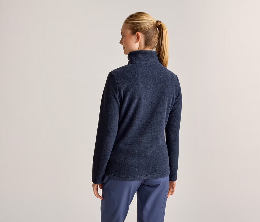 Eine Frau von hinten, die eine dunkelblaue Fleecejacke und eine blaue Hose trägt. Ihr blondes Haar ist zu einem Pferdeschwanz gebunden.