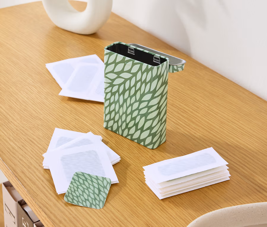 Auf einem Holztisch stehen eine Dose mit grünem Blattmuster, leere Wachsstreifen und eine Vase mit Pflanzen.