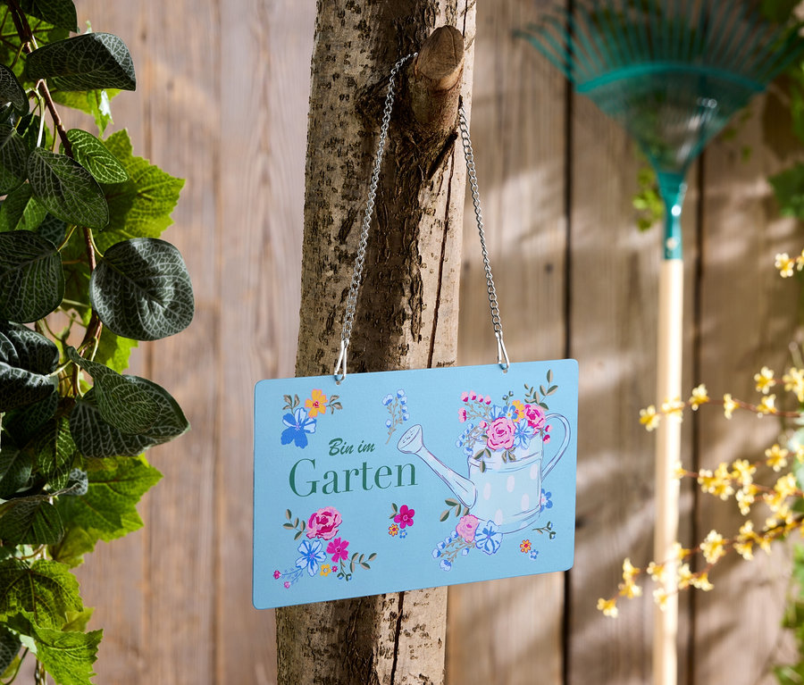 Ein blaues Schild mit der Aufschrift "Bin im Garten" hängt an einem Baum, im Hintergrund ein Rechen.