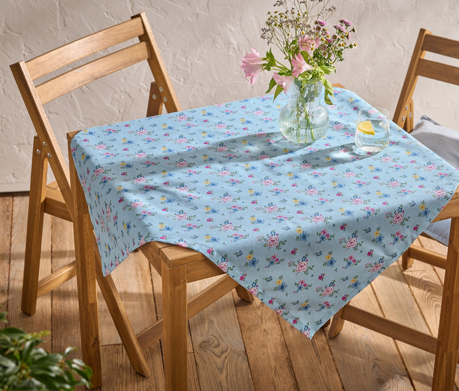 Tisch mit hellblauer Tischdecke mit Blumenmuster, Vase mit Blumen und Glas Wasser mit Zitrone auf zwei Holzstühlen.