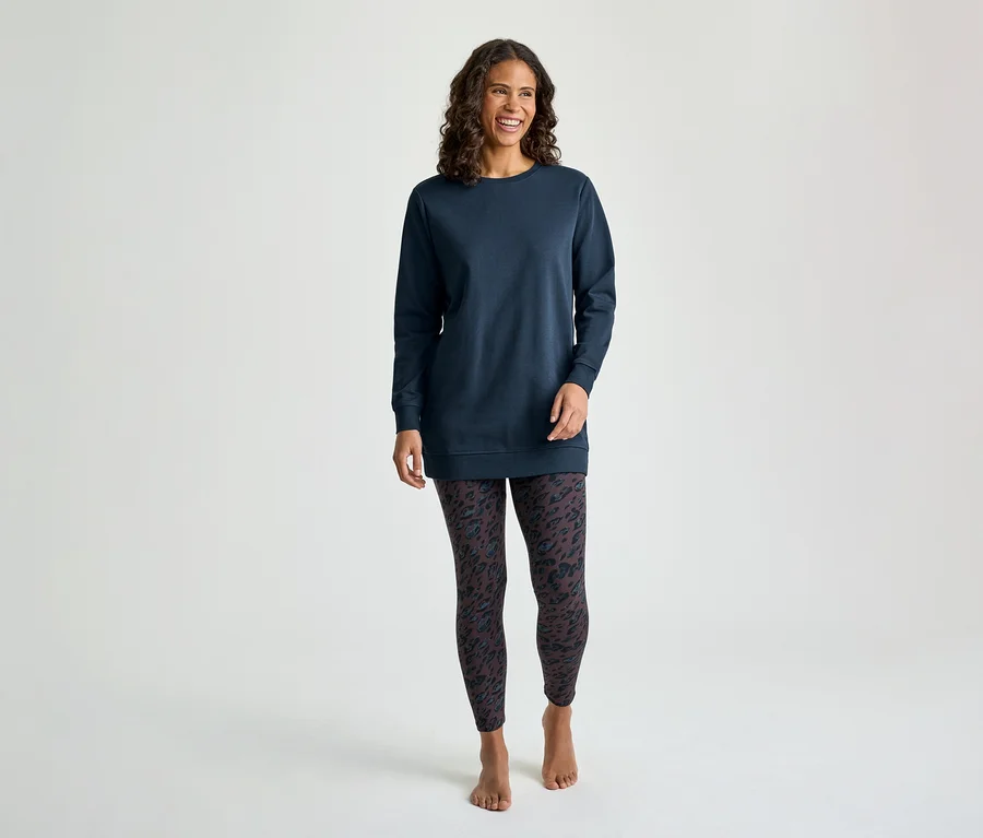 Eine lächelnde Frau mit lockigem braunem Haar steht frontal und trägt ein langärmeliges, marineblaues Tunika-Oberteil und dunkle gemusterte Leggings. Sie ist barfuß.