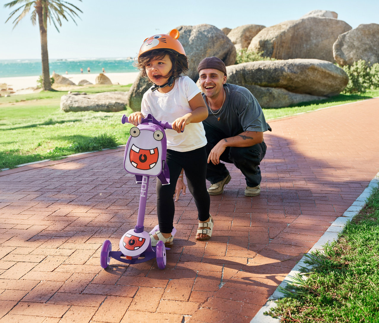 Kind mit orangefarbenem Helm fährt Hudora 4-in-1-Scooter, Lavendel, auf einem Ziegelweg. Ein Mann kauert daneben.