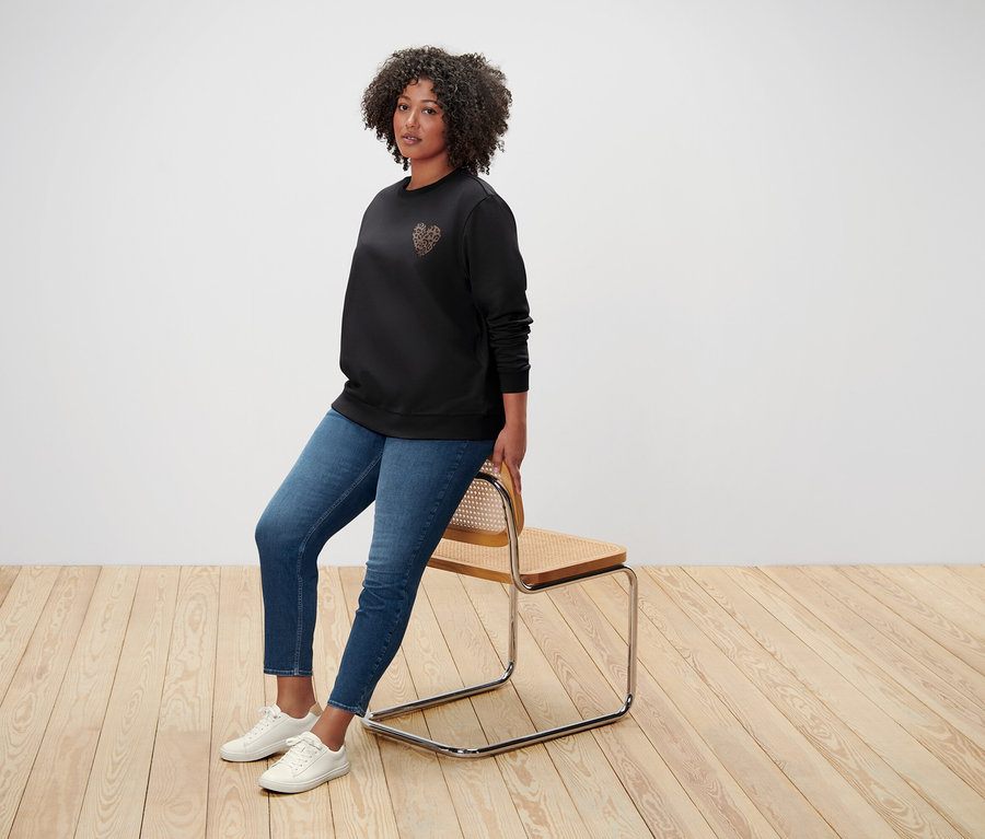 Eine Frau mit lockigem Haar sitzt auf einem Stuhl, gekleidet in einen schwarzen Pullover mit Herzmotiv, Jeans und weißen Turnschuhen.