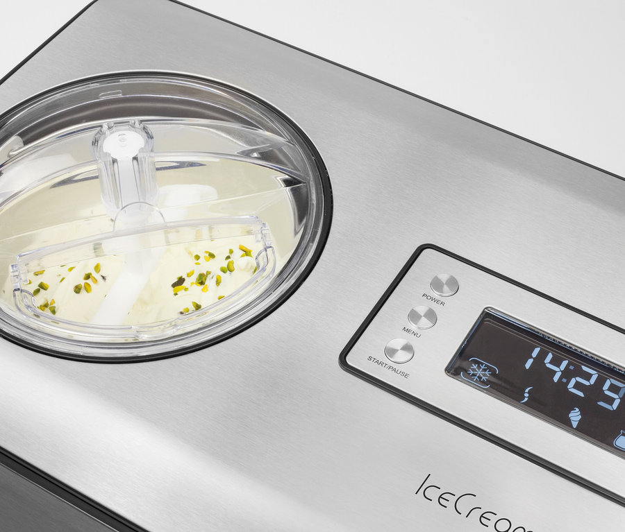Eine Eismaschine aus gebürstetem Edelstahl mit einem transparenten Deckel, unter dem hellgelbes Eis mit Pistazienstücken gerührt wird. Das digitale Display zeigt "14:29" an.