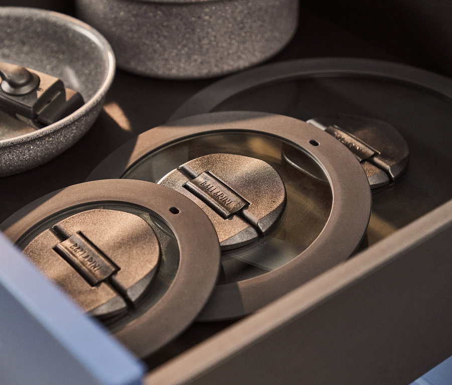 Eine Küchenschublade enthält gesprenkeltes graues Kochgeschirr: eine Bratpfanne mit abnehmbarem Griff und zwei runde, metallische Deckel mit der Prägung 'BALLARINI'.