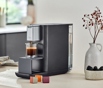 Qbo ESSENTIAL Dark Stone Kaffeemaschine füllt ein Glas mit Kaffee. Daneben stehen Qbo ICONIC.