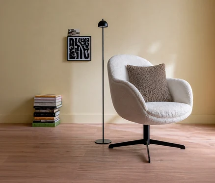 Ein weißer Sessel mit einem taupefarbenen SCHÖNER WOHNEN-Kollektion Dekokissenbezug »TEDDY« steht auf einem Holzboden. Daneben eine schwarze Stehlampe, im Hintergrund eine gelbe Wand mit einem gerahmten Bild und einem Stapel Bücher.