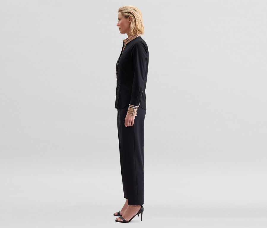 Frau im Profil trägt eine schwarze Seidensticker Popeline-Bluse mit Karodetail an Kragen und Manschetten, schwarze Hose und schwarze High Heels.
