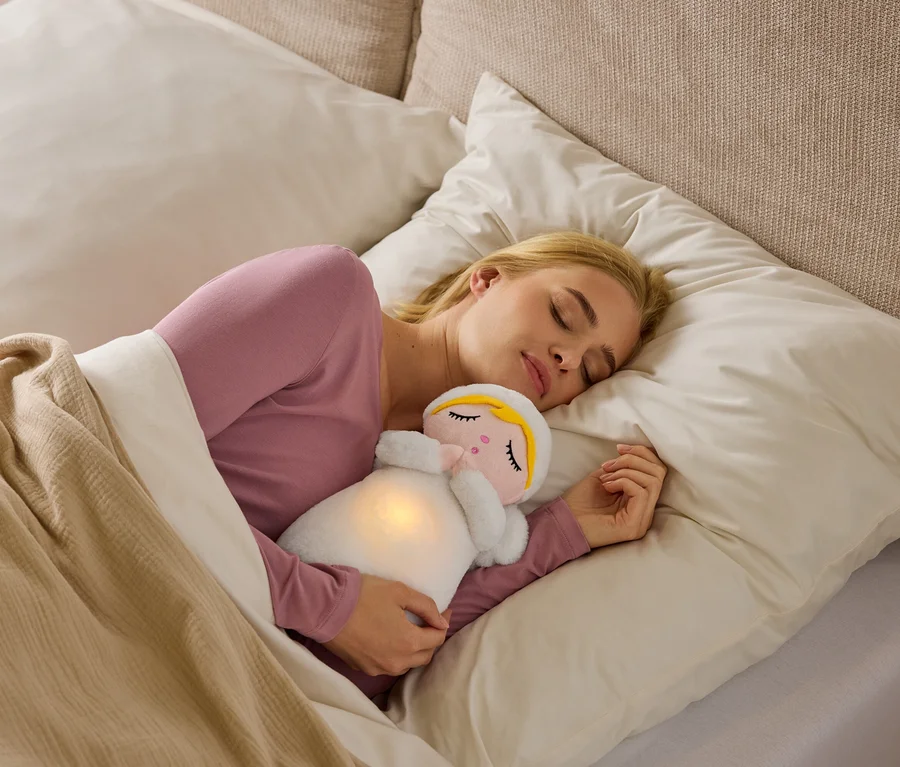Eine blonde Frau schläft auf der Seite im Bett, trägt ein rosa Oberteil und kuschelt ein weißes Plüschtier mit leuchtendem Bauch unter einer beigen Decke.