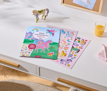 Weißer Schreibtisch mit einem Stickerbuch „MEIN STICKERBUCH Einhörner“ (über 350 Sticker), drei Stickerbögen, einer Einhornfigur, einem Glas Orangensaft, einem rosa Plüsch-Notizbuch, einem Einhornstift und einem Einhornarmband.