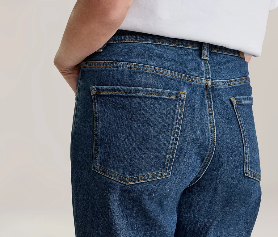 Person von hinten in blauen Jeans und weißem T-Shirt. Die Jeans hat sichtbare Gesäßtaschen und gelbe Nähte.