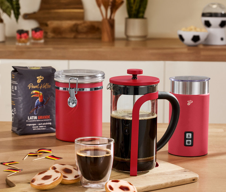 Auf einem Tisch stehen eine rote Aromadose, ein roter, elektrischer Milchaufschäumer, eine rote Siebstempelkanne, 2 Caffè Crema Gläser und eine Kaffeepackung.