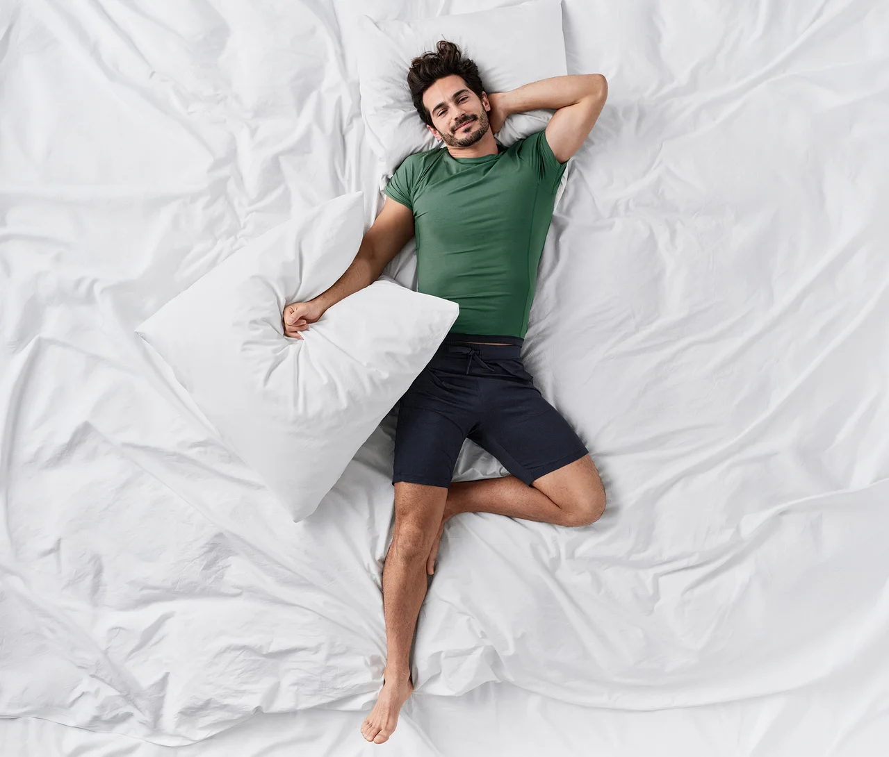 Ein lächelnder Mann liegt auf dem Rücken in einem Bett mit weißen Laken und trägt ein grünes CALIDA »Deepsleepwear Balancing« T-Shirt und dunkle CALIDA »Deepsleepwear Balancing« Shorts.