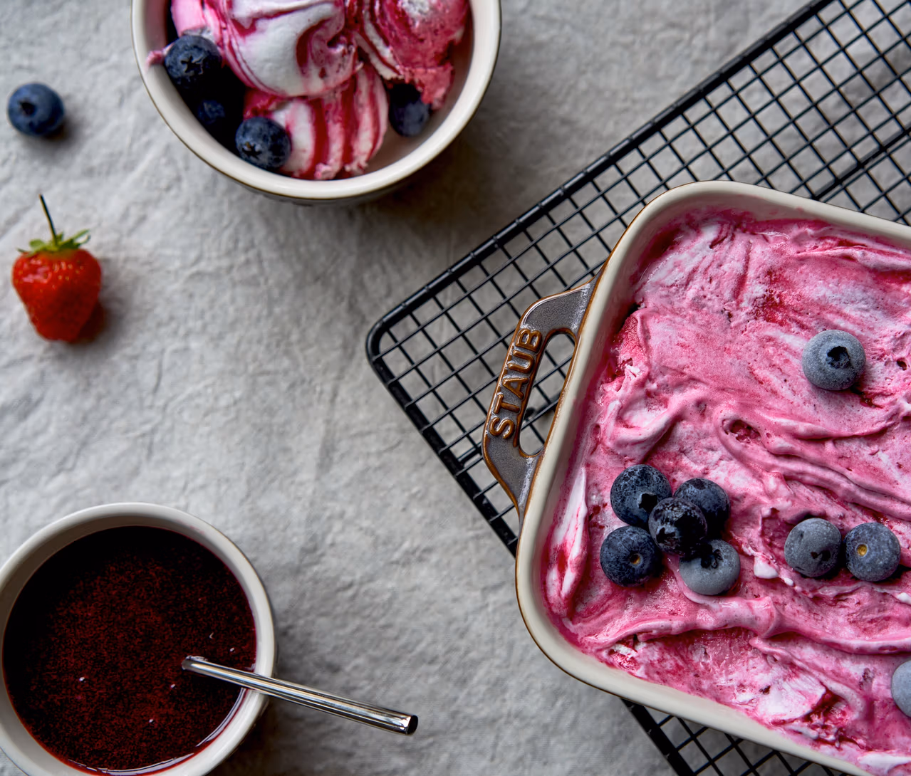 Blick von oben auf eine rechteckige, antikgraue STAUB Auflaufform mit pinkfarbenem gefrorenem Dessert und Heidelbeeren, daneben eine Schale mit Beeren-Eis, eine Schale mit Beerensauce und Löffel sowie eine Erdbeere auf hellgrauem Untergrund.