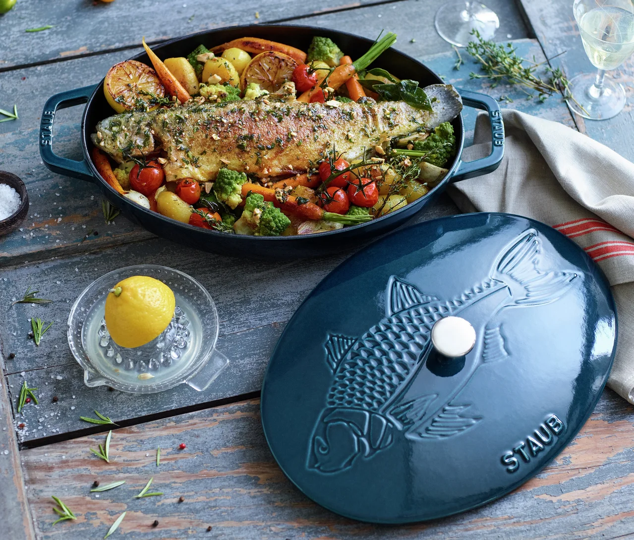 Gebratener Fisch mit Gemüse in einer ovalen blauen Staub-Auflaufform, daneben ein passender Deckel mit Fischmotiv und eine Zitrone auf einer Saftpresse.
