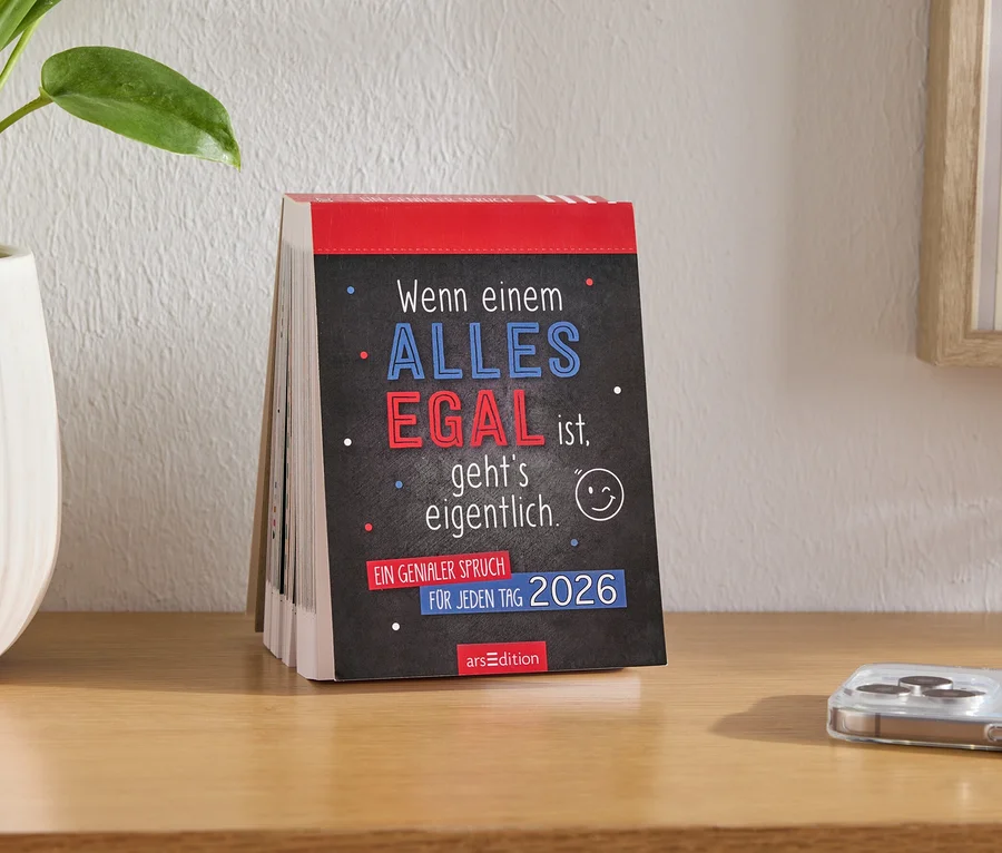 Tischkalender mit der Aufschrift "Wenn einem ALLES EGAL ist, geht's eigentlich 2026", neben Blumentopf und Mobiltelefon auf dem Tisch.