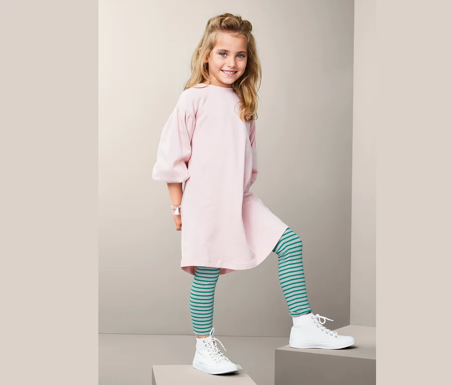 Mädchen posiert auf Stufen, trägt ein rosa Kleid und 2 Leggings mit blaugrünen Streifen.