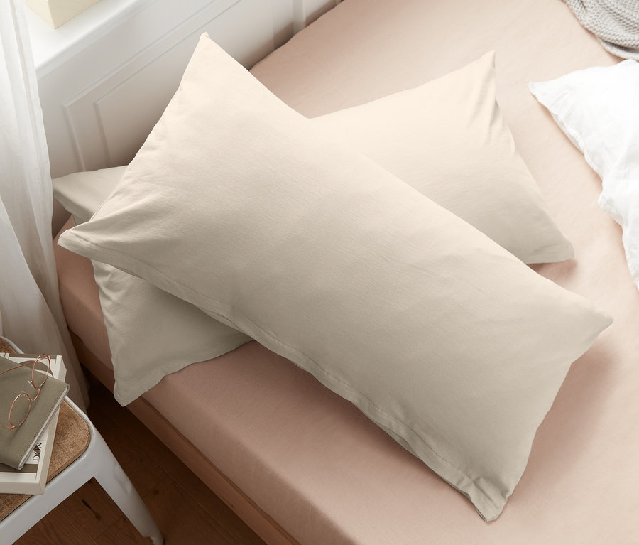 Zwei cremefarbene Jersey-Kopfkissenbezüge liegen auf einem Bett neben Büchern und einer Brille.