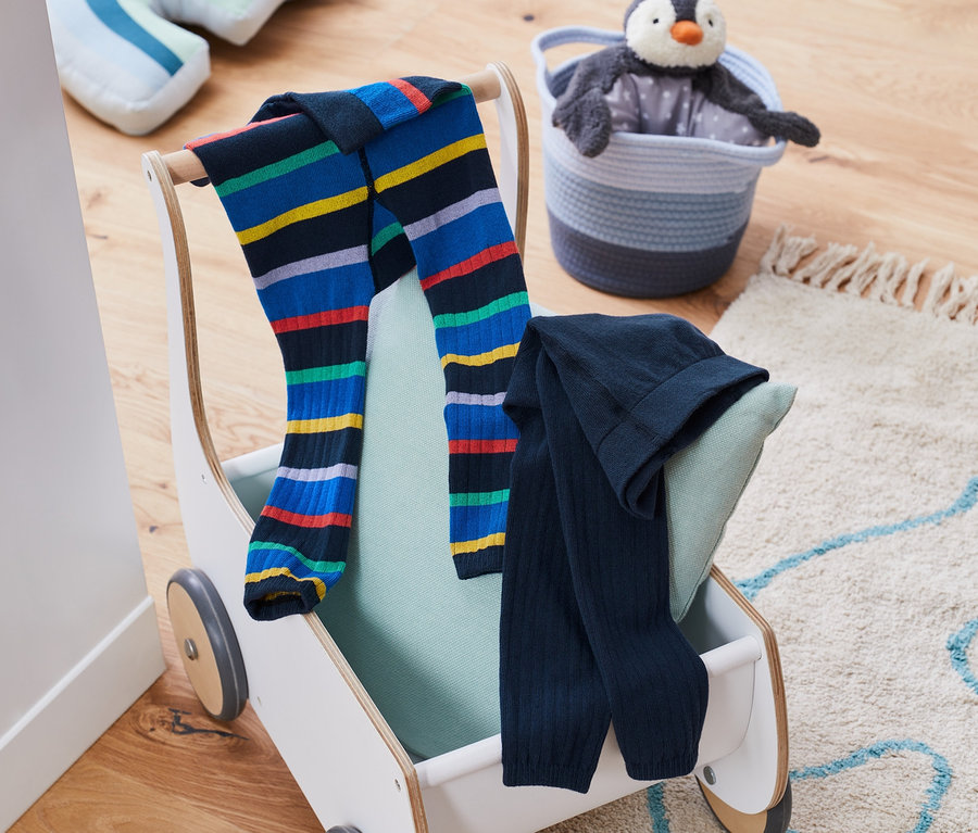 Zwei Strickleggings liegen auf einem Spielzeugwagen neben einem Plüschpinguin in einem Korb.