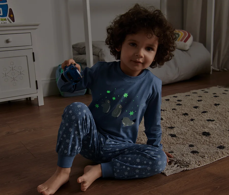 Ein Junge sitzt auf dem Boden und hält ein blaues Spielzeugauto in der Hand. Er trägt einen Kinder-Glow-in-the-dark-Pyjama mit Pinguinprint.