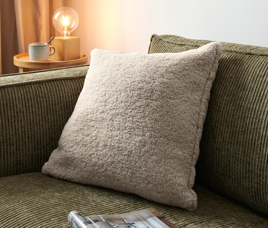 Ein beiges Dekokissen liegt auf einem olivgrünen Sofa neben einer offenen Zeitschrift.