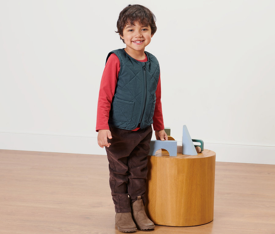 Ein Junge steht neben einem Holztisch und trägt eine Kinder-Wendesteppweste.