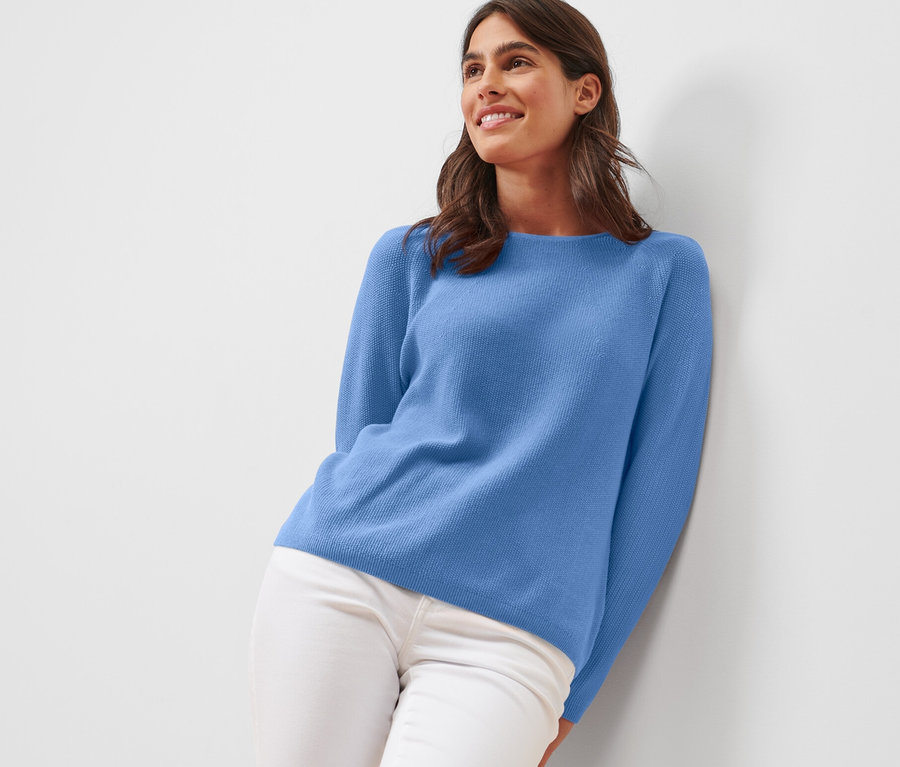 Frau an eine Wand gelehnt, trägt einen blauen Strickpullover.