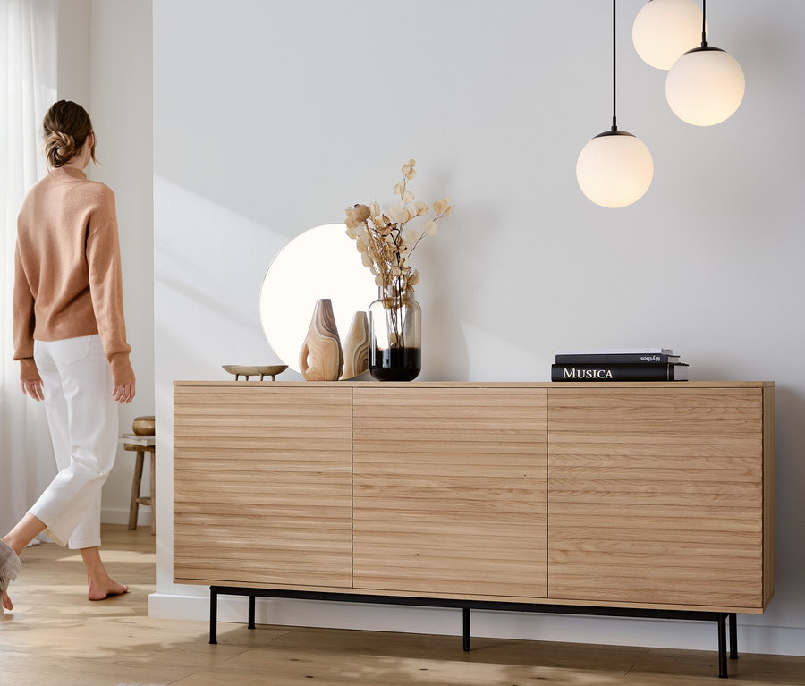 Eine Frau geht an einem Sideboard »Finnsbo« mit Lamellenoptik vorbei, über dem eine 3-flammige Hängeleuchte mit Glaskugeln hängt. Sessel mit Drehfunktion im Vordergrund.