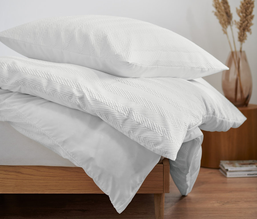Drei Sets Struktur-Bettwäsche in Normalgröße liegen auf einem Bett. Im Hintergrund befinden sich ein runder Spiegel und eine Vase.