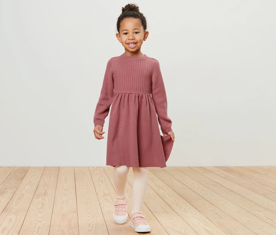 Ein lächelndes Mädchen geht auf einem Holzboden und trägt ein Kinder-Merinostrickkleid.