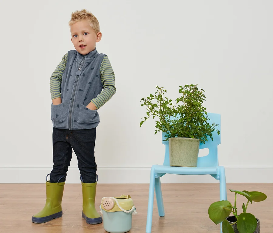 Junge steht mit Händen in den Taschen in Kleinkinder-Fleeceweste, Schnecken-Alloverprint, Jeans und Gummistiefeln.
