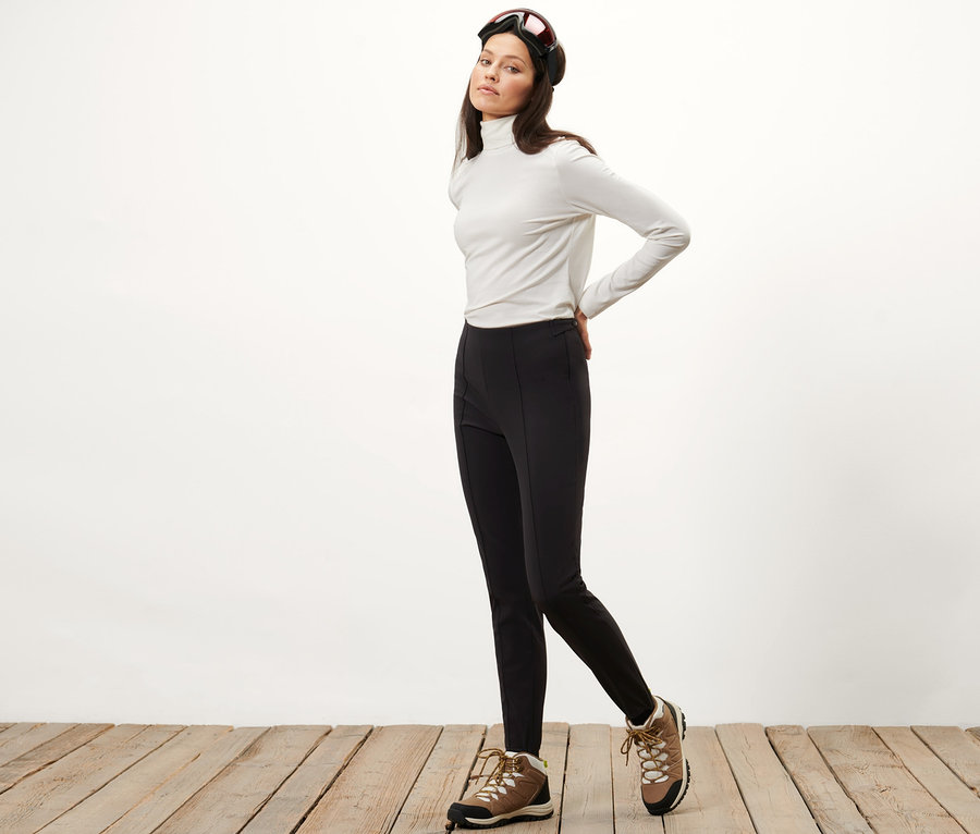 Frau posiert in Softshell-Ski-Tight, weißem Rollkragenpullover und Skibrille.