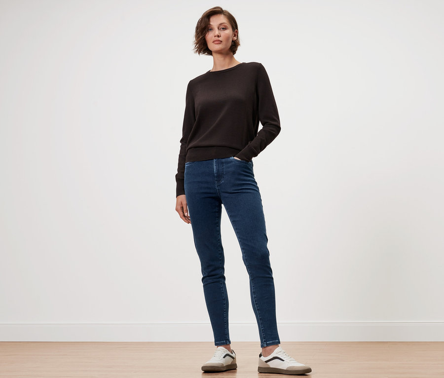 Frau posiert in braunem Merino-Feinstrickpullover, blauen Jeans und Sneakers.