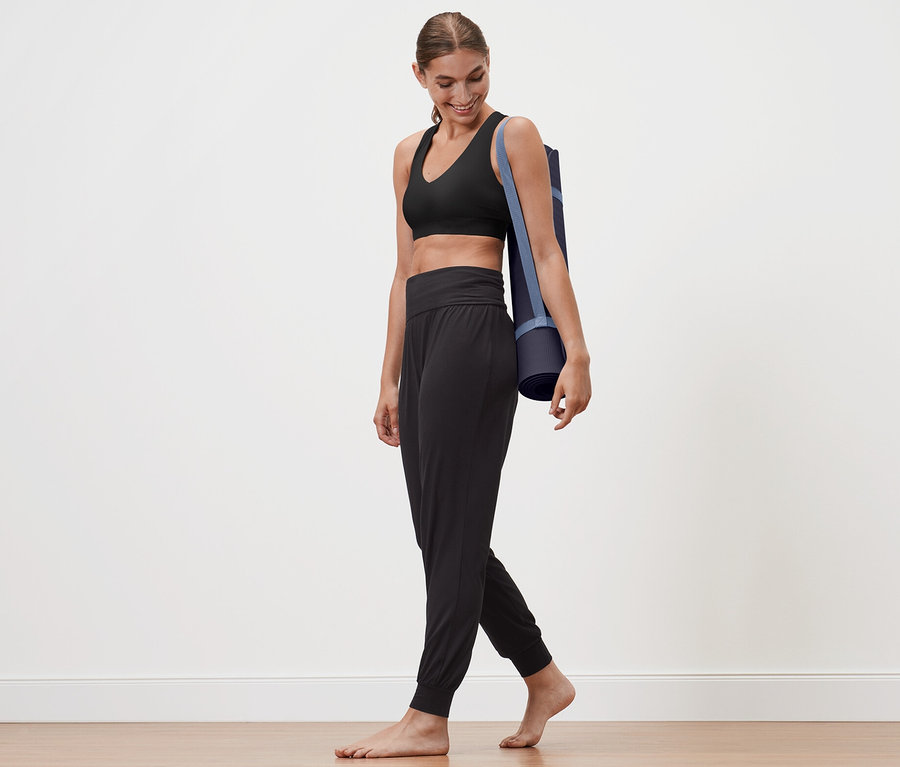 Frau geht mit einer Yogamatte über der Schulter. Sie trägt eine Yogahose.