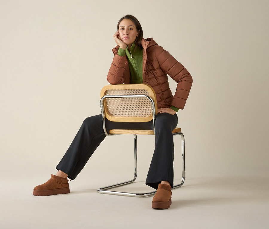 Frau sitzt auf einem Stuhl mit Strick-Troyer, Jacke und Schuhen.