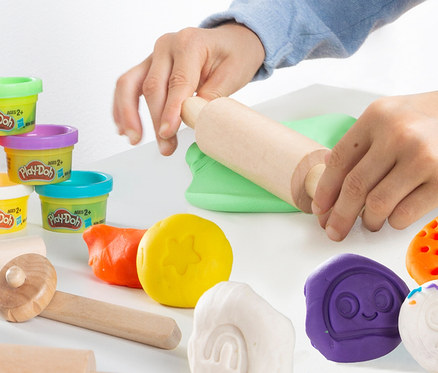 Kinderhände rollen grüne Knete mit einer Walze aus dem roba x Play-Doh Knet-Spielset 1. Im Vordergrund stehen Becher und weitere Knete.