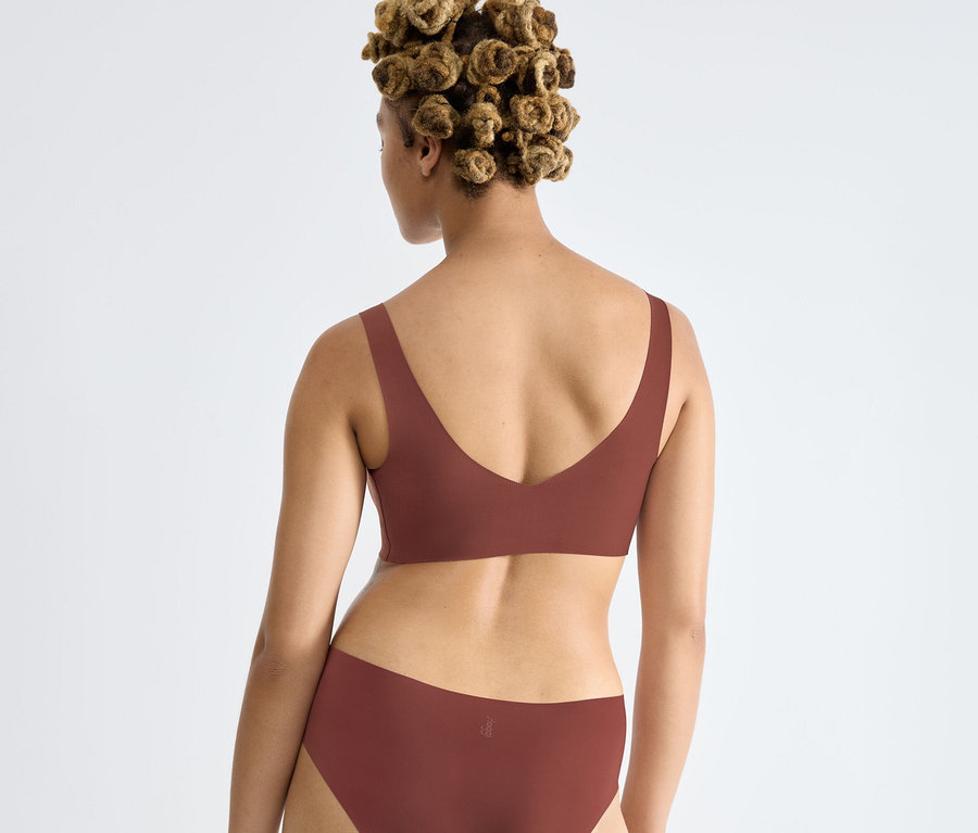 Frau von hinten in einem braunen sloggi ZERO Feel 2.0 Bralette und passender Unterhose, mit lockigem Haar zu kleinen Knoten gebunden.